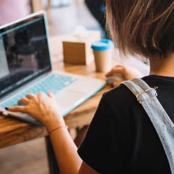 picture looking over the shoulder of a woman wearing a black shirt working at a laptop on a wooden desk